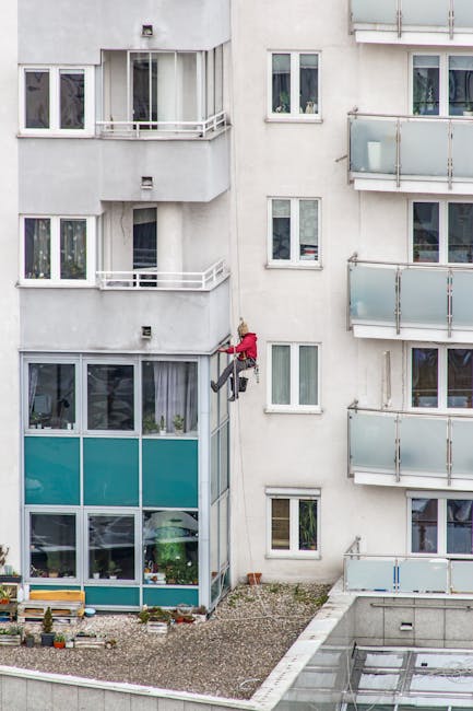 Professional window cleaner scaling a modern residential building with glass balconies.