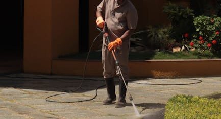 A gardener wearing a hat cleans an urban sidewalk with a pressure washer.